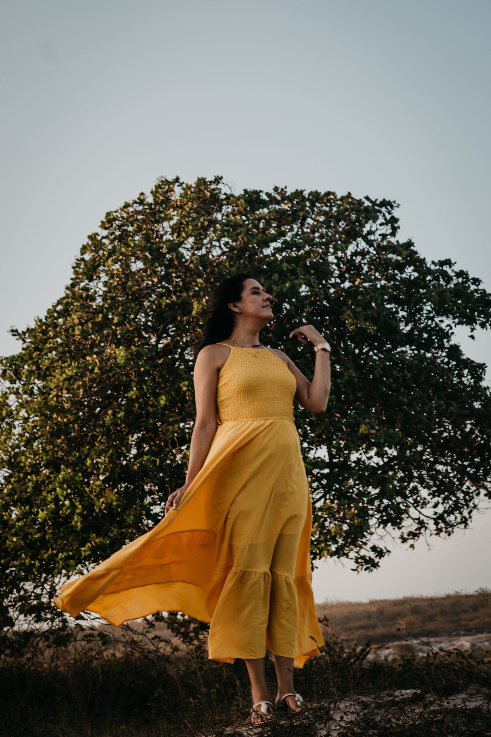 Elegant woman in yellow dress stands in a summer park, enjoying a serene moment with a gentle breeze.