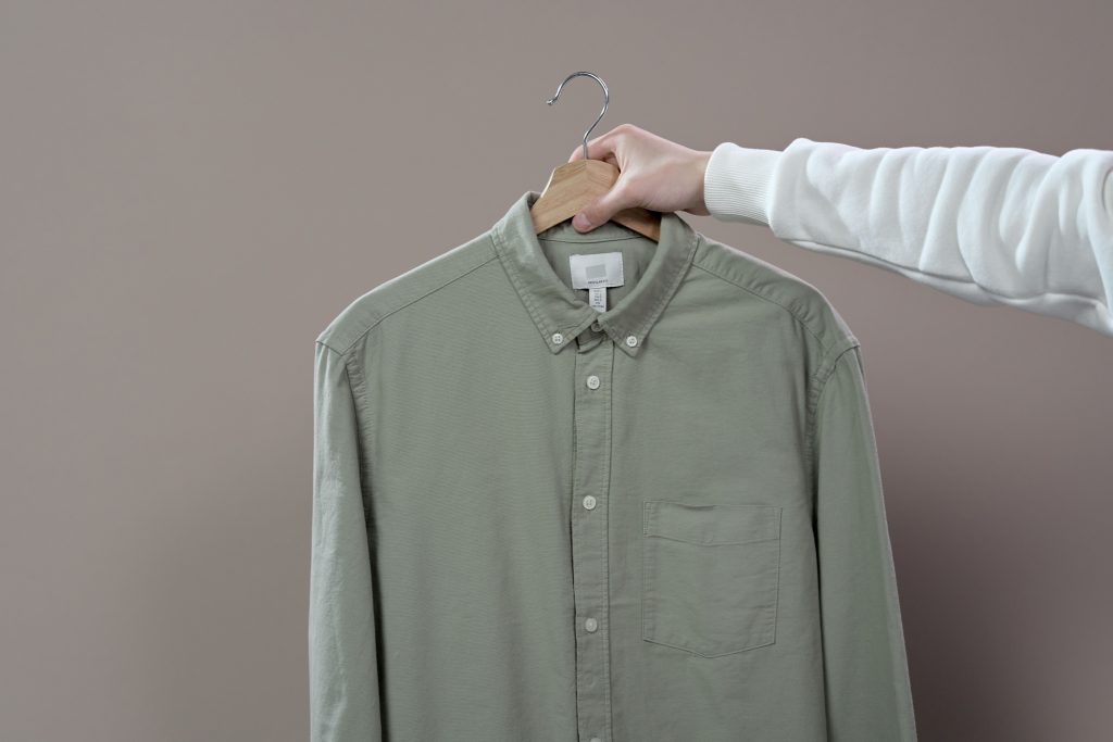 A hand holding a green button-up shirt on a hanger in a minimalist studio setting.
